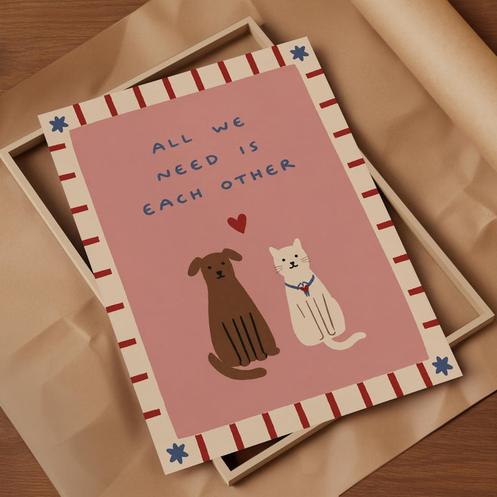 a card with a pink background and a border of red and white stripes. The card has a message that reads "All we need is each other" and includes illustrations of a brown dog and a white cat.