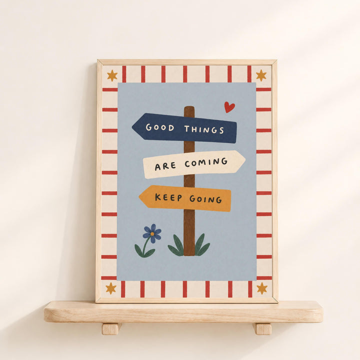 The image is a framed poster with a blue background and a border of red and white stripes. The poster features a signpost with three directional signs, each with a different color and text. The signpost reads "GOOD THINGS ARE COMING KEEP GOING".
