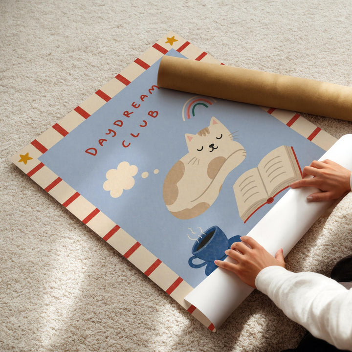 A child's hands are seen rolling up a poster with a cat illustration and other playful elements, such as a cup and a book, on a carpeted floor.