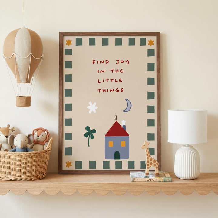 a framed poster with a whimsical illustration of a house, a giraffe, and a hot air balloon, along with the text "Find joy in the little things". The poster is displayed on a shelf, accompanied by a basket of stuffed animals and a lamp.