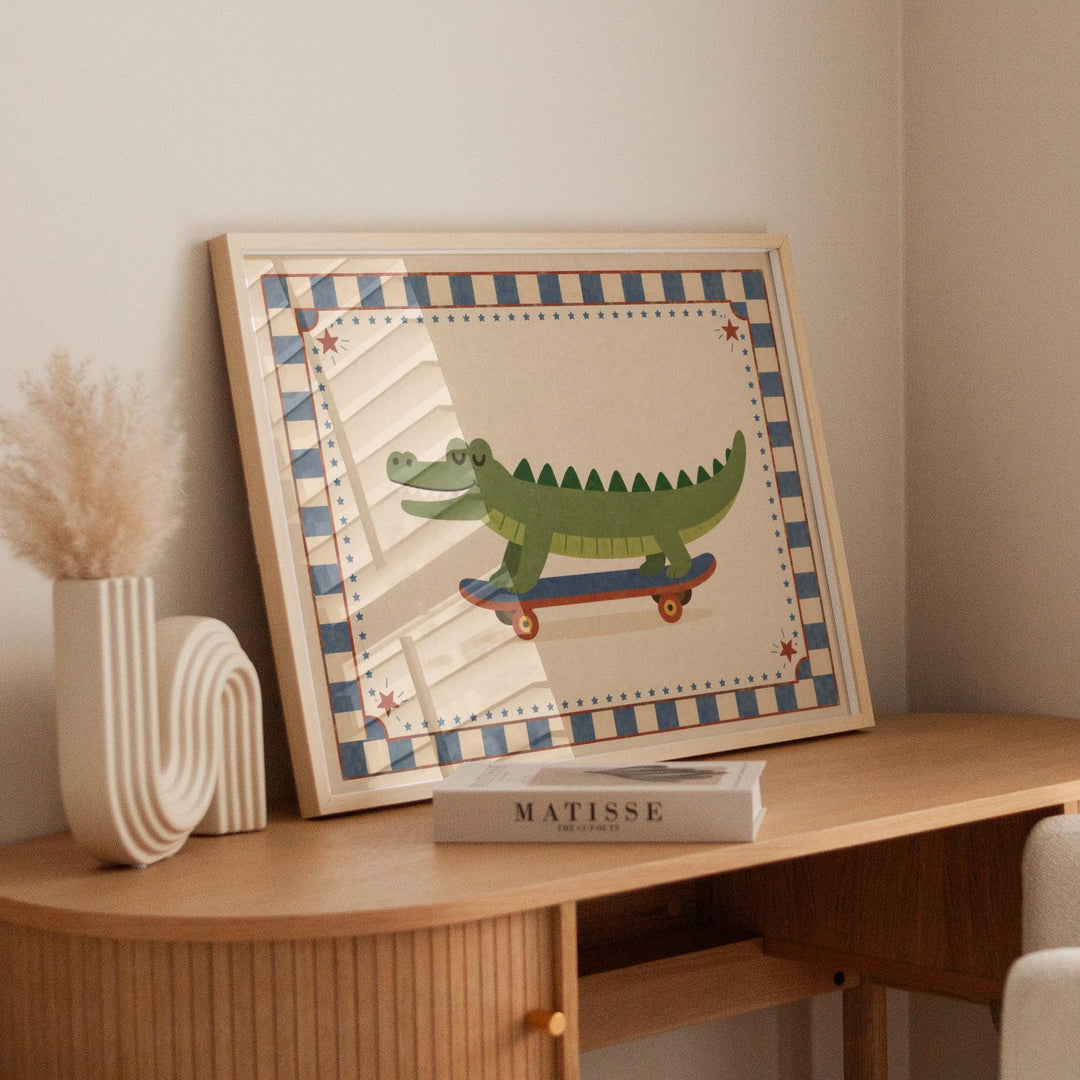 a framed artwork of a cartoon alligator riding a skateboard, placed on a wooden table. Next to the artwork, there is a book titled "MATISSÉ" and a white vase.