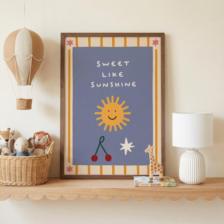 a framed poster with a cheerful design, including a sun, cherries, and a giraffe, along with the text "Sweet like sunshine". The poster is displayed on a shelf, accompanied by a basket of stuffed animals and a lamp.