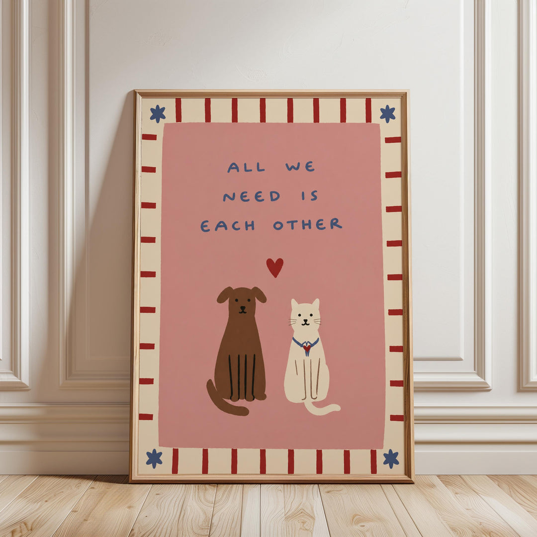 A framed poster with a pink background and a border of red and white stripes. The poster features two cartoon animals, a dog and a cat, sitting together with a heart symbol between them. The text on the poster reads "All we need is each other".