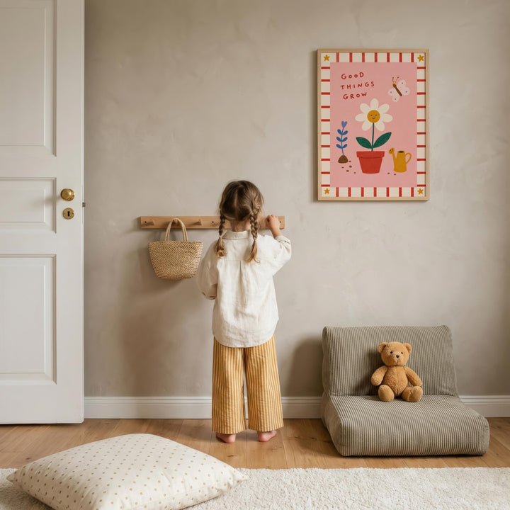 A young girl stands in a cozy room, looking at a framed picture on the wall. The room has a wooden floor, a small chair, and a teddy bear on the floor.