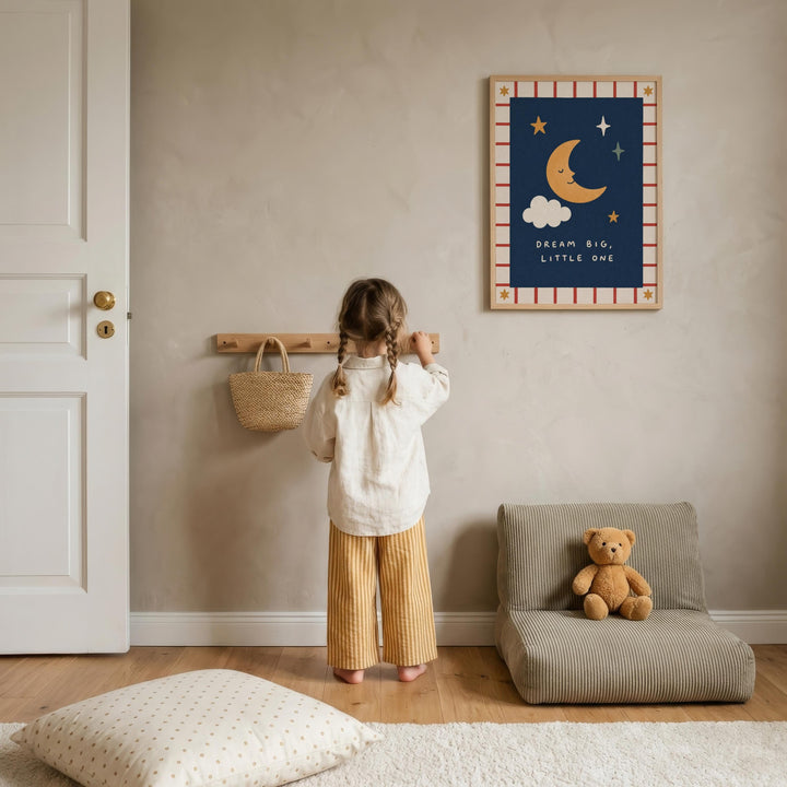 A young girl stands in a cozy room, looking up at a framed picture on the wall. The room has a wooden floor, a chair, and a teddy bear on the floor.