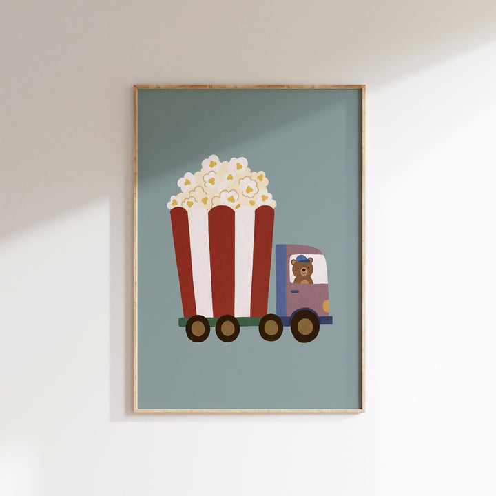 A framed illustration of a red and white striped truck carrying a large bucket of popcorn, with a teddy bear inside the truck.