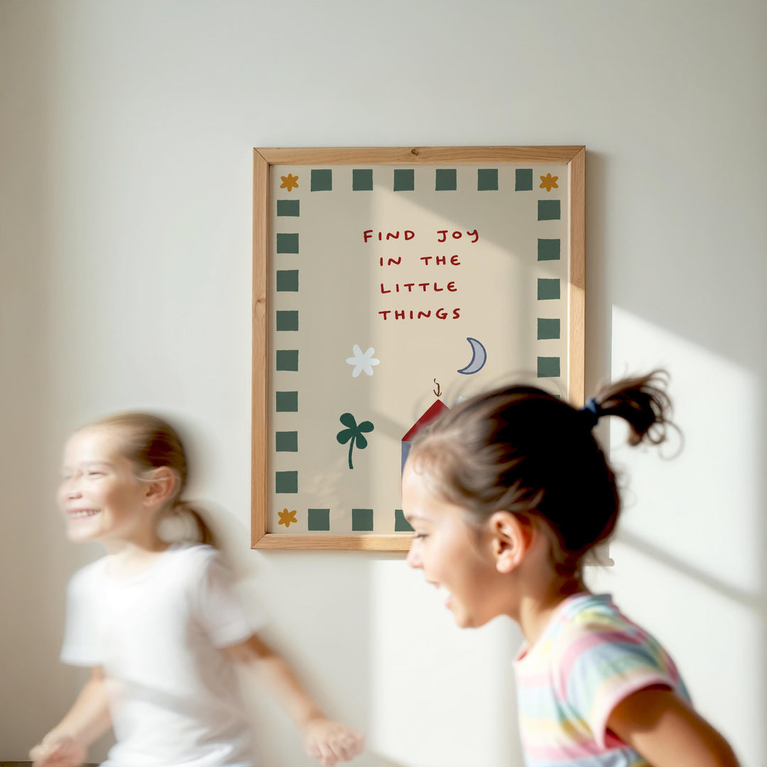 Two young girls are running in front of a framed poster with the text "Find joy in the little things" on a wall.
