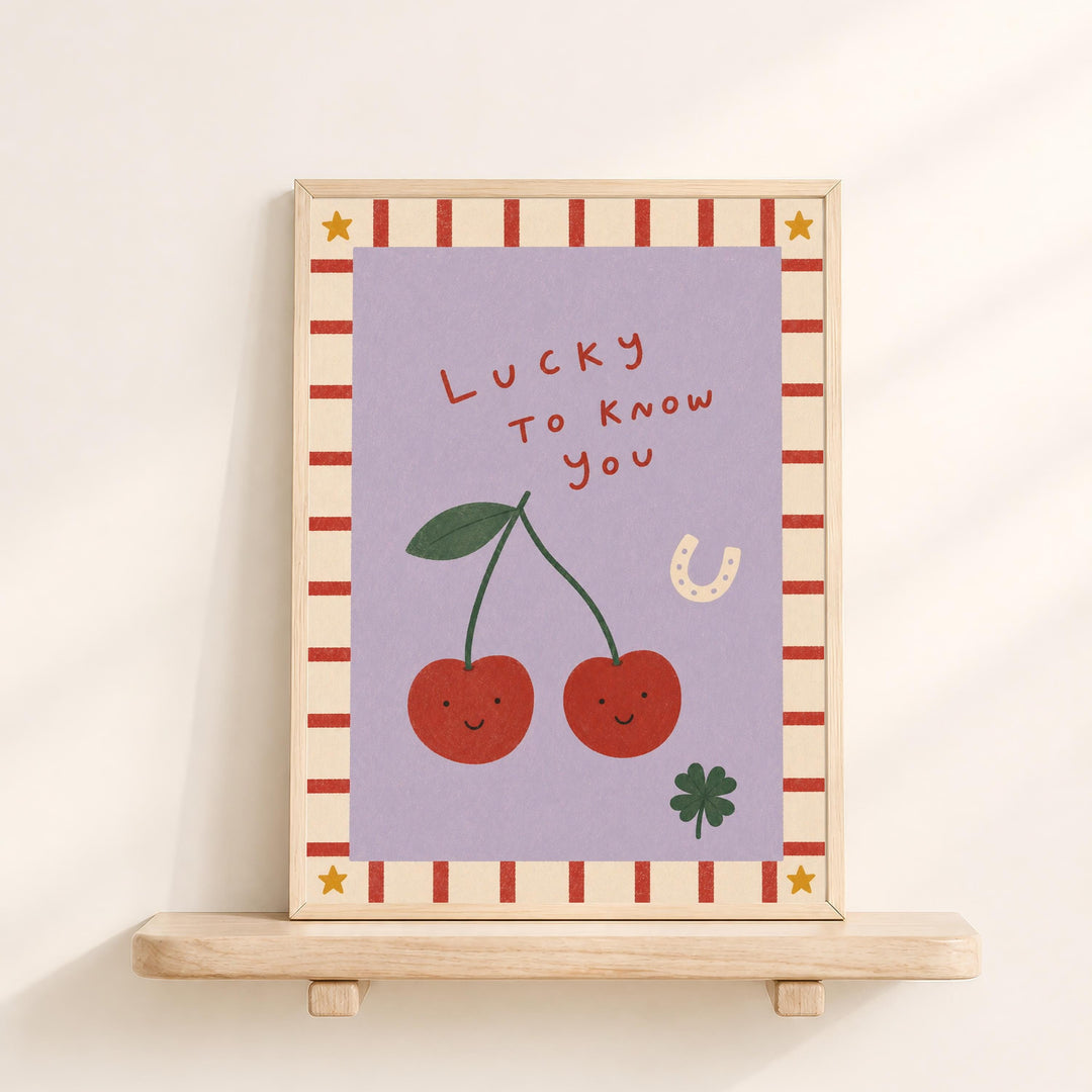 The image is a framed illustration featuring two cherries with smiling faces, a horseshoe, and a clover, all set against a purple background with a striped border.