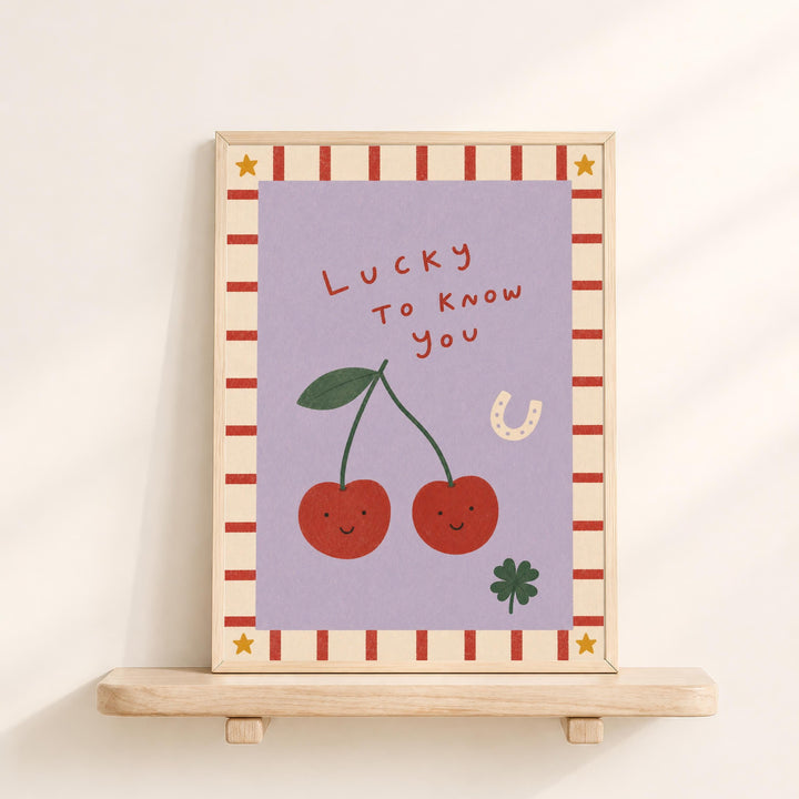 The image is a framed illustration featuring two cherries with smiling faces, a horseshoe, and a clover, all set against a purple background with a striped border.