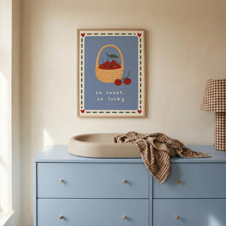 a blue dresser with a white basin, a framed poster of cherries on the wall above it, and a plaid blanket on the dresser.