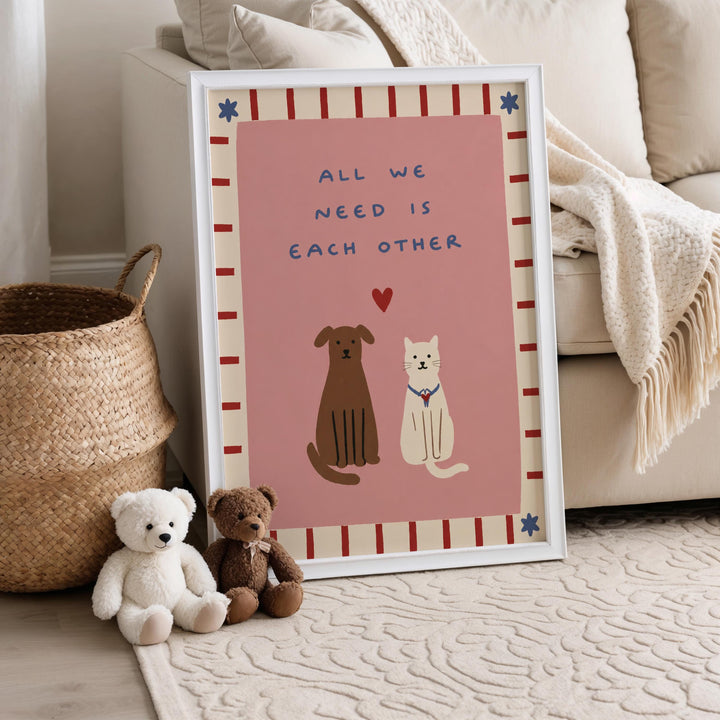 A framed poster with a pink background and a message about the importance of companionship between dogs and cats. The poster is placed on a carpeted floor next to a white basket and a stuffed teddy bear.