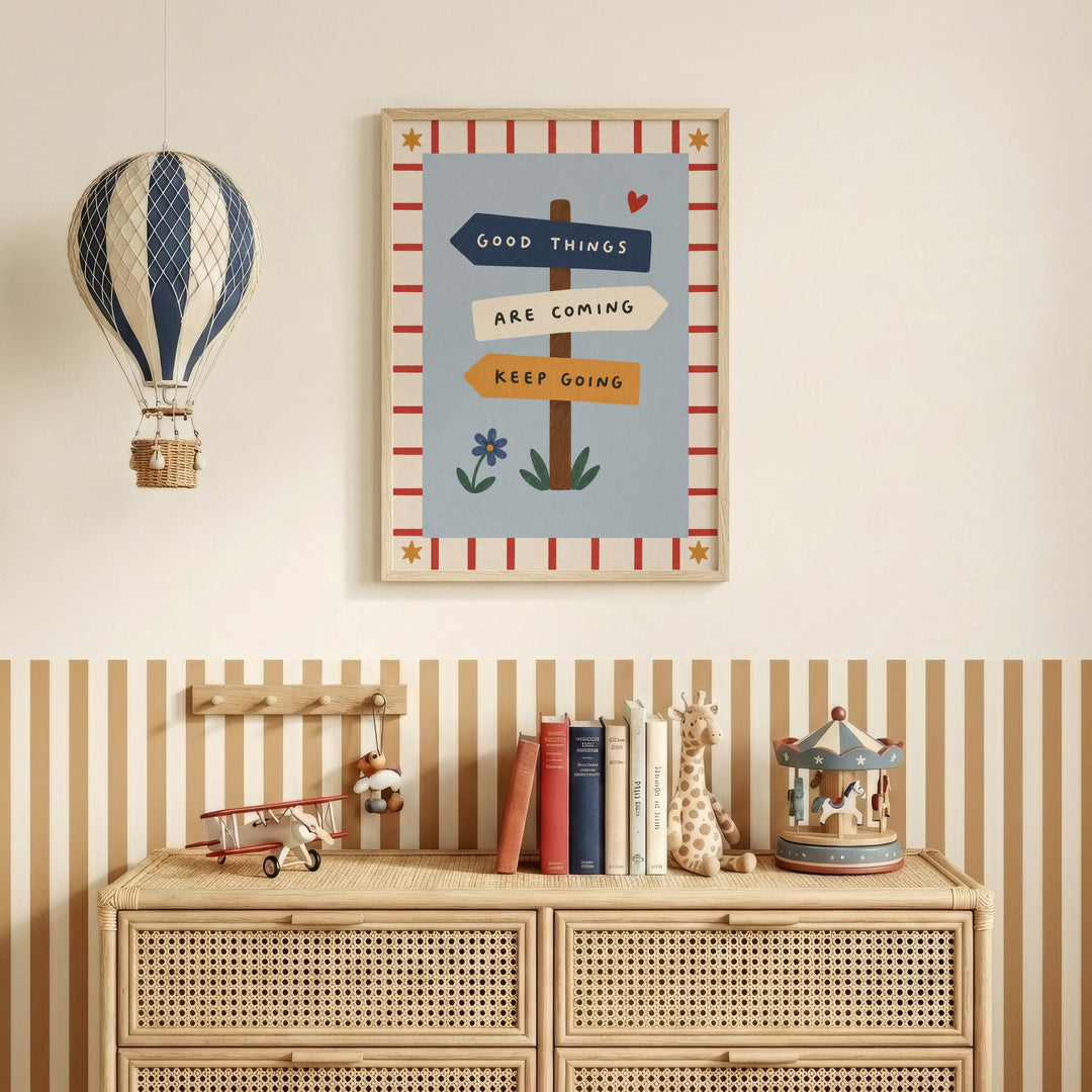 a wall with a framed poster that says "GOOD THINGS ARE COMING KEEP GOING" and a hot air balloon hanging above it. Below the poster, there is a wooden dresser with a toy airplane, a carousel, and a collection of books.