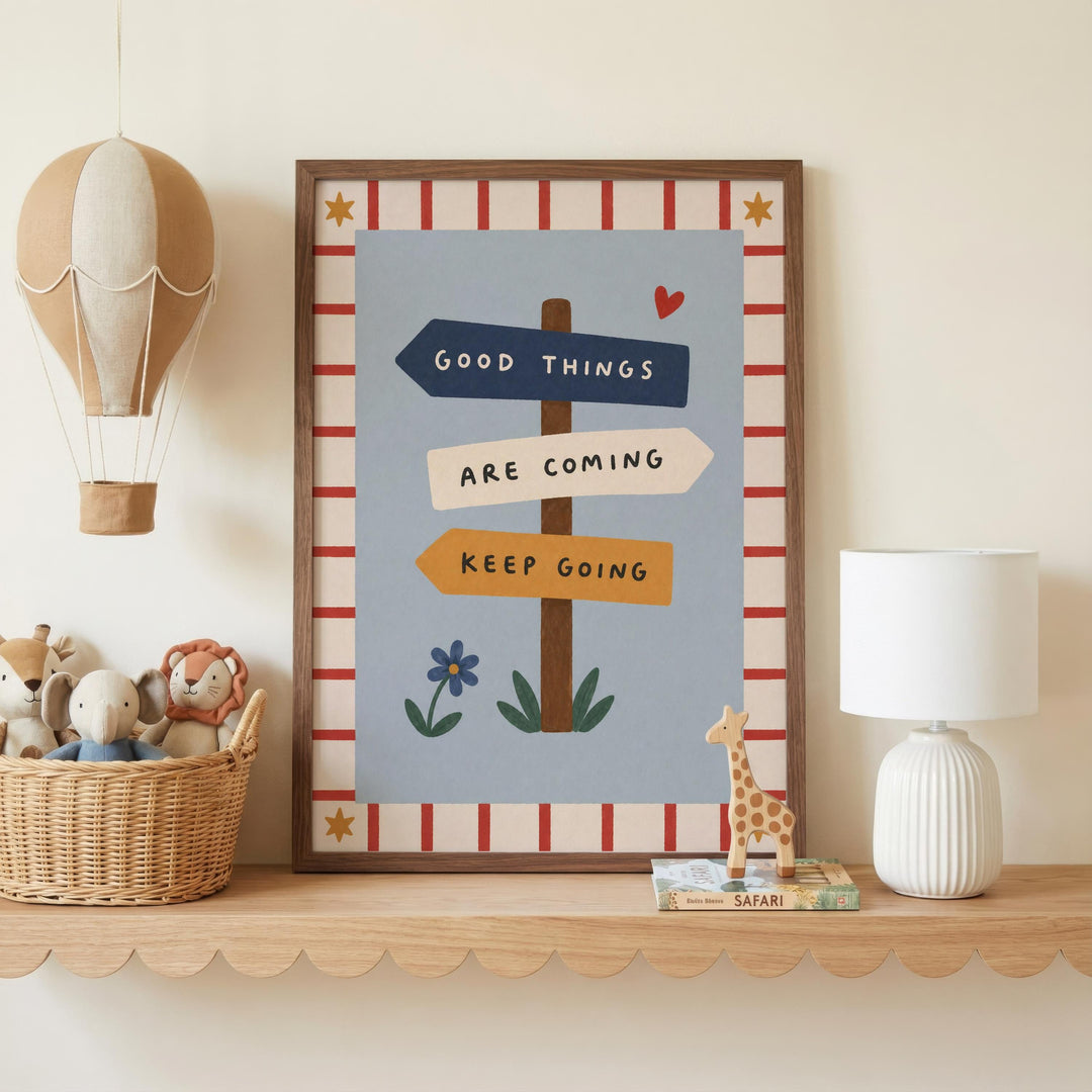 a framed poster with the text "GOOD THINGS ARE COMING KEEP GOING" and illustrations of a hot air balloon, a heart, a giraffe, and a flower. The poster is displayed on a shelf, accompanied by a lamp, a basket of stuffed animals, and a small toy giraffe.