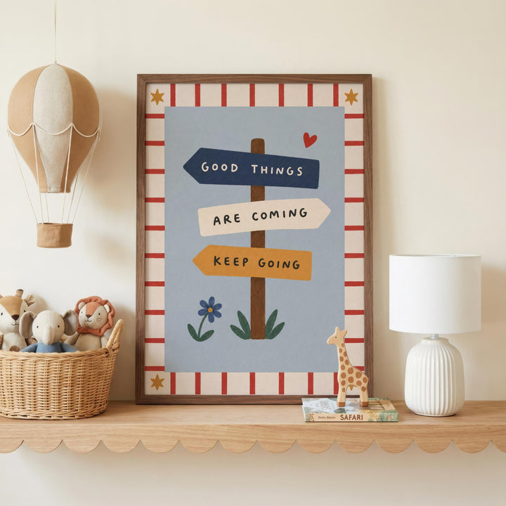 a framed poster with the text "GOOD THINGS ARE COMING KEEP GOING" and illustrations of a hot air balloon, a heart, a giraffe, and a flower. The poster is displayed on a shelf, accompanied by a lamp, a basket of stuffed animals, and a small toy giraffe.