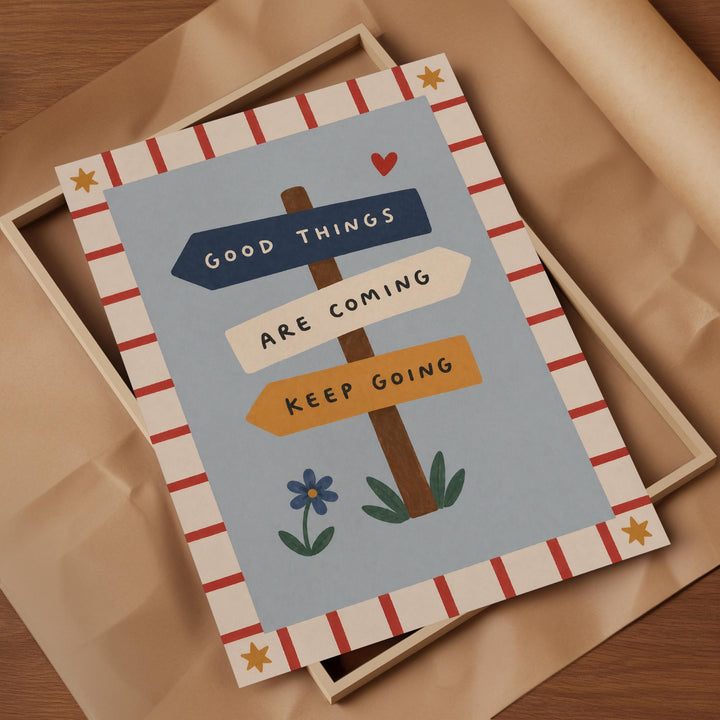 The image is a framed piece of art with a blue background and a wooden post with three directional signs. The text on the sign reads "Good Things Are Coming" and "Keep Going".