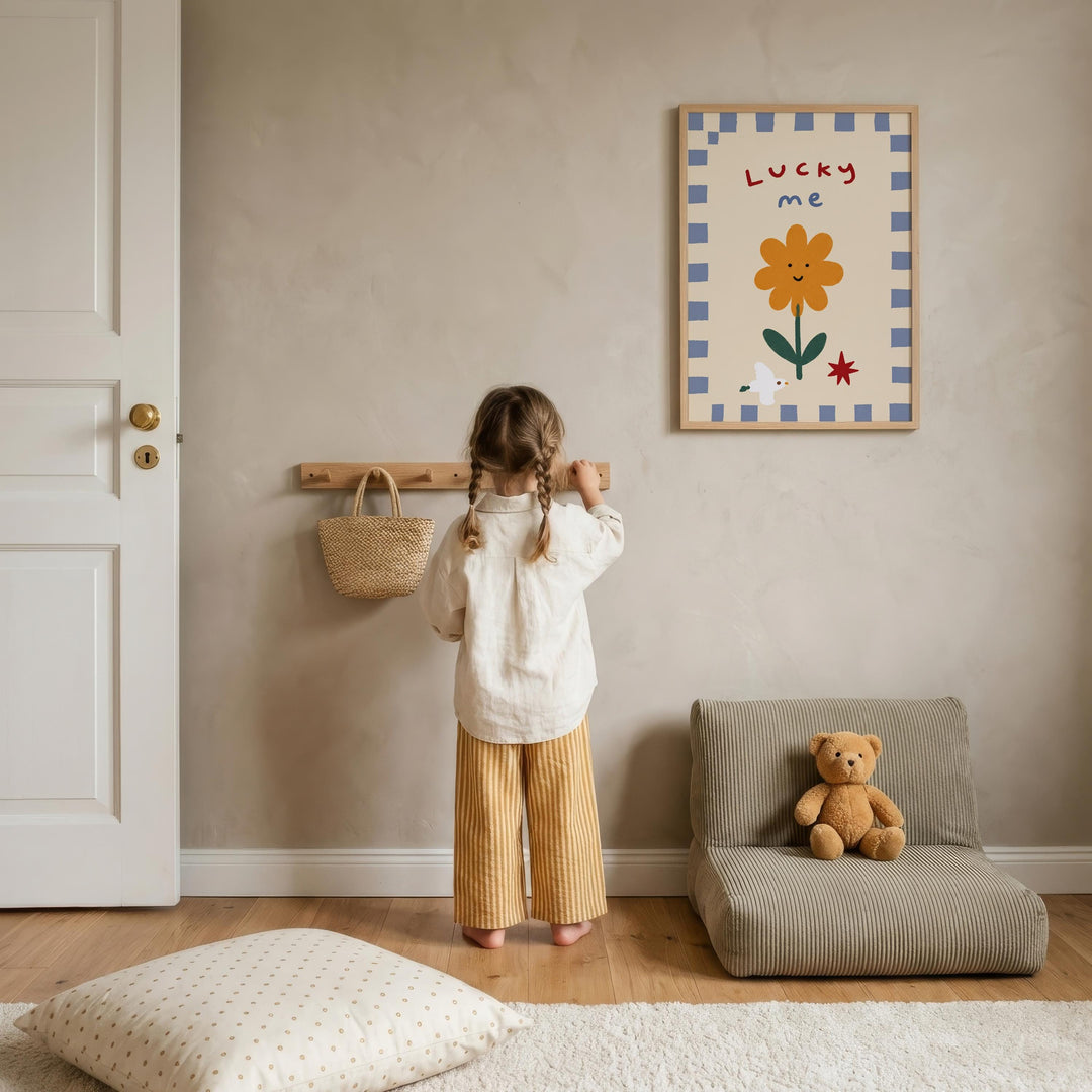 A young girl stands in a cozy room, looking at a framed picture on the wall that says "Lucky me". The room has a wooden floor, a small chair, and a teddy bear on the floor.