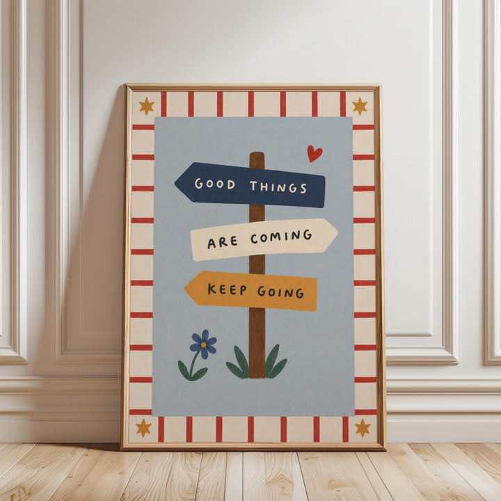 A framed poster with a blue background and a red and white border, featuring a sign with the text "GOOD THINGS ARE COMING KEEP GOING" and a small blue flower.