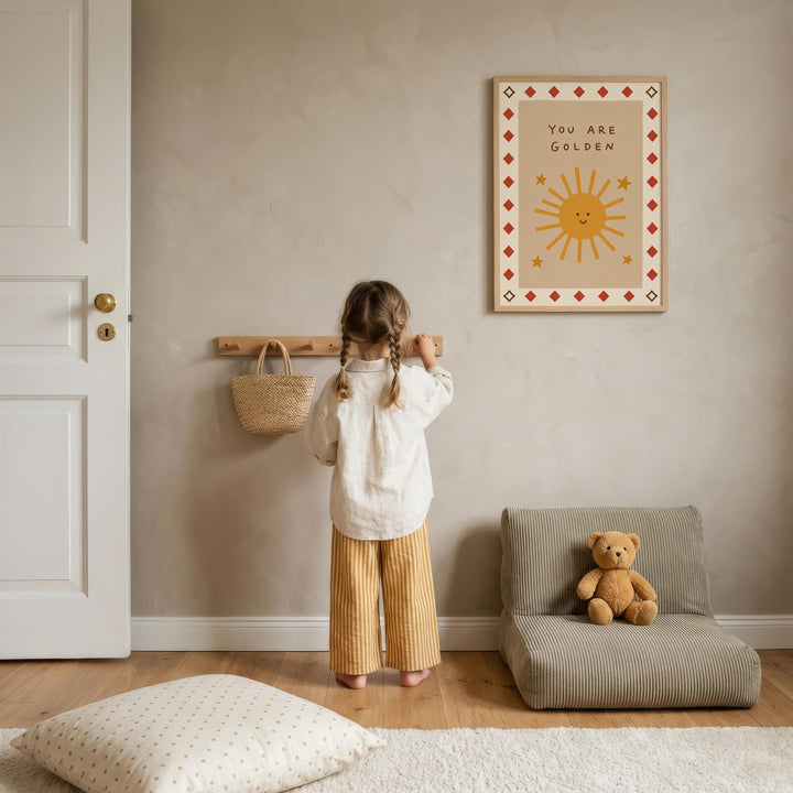 A young girl stands in a cozy room, looking at a framed poster with a sun and stars design. The room has a beige color scheme, with a teddy bear on a chair and a basket on the wall.