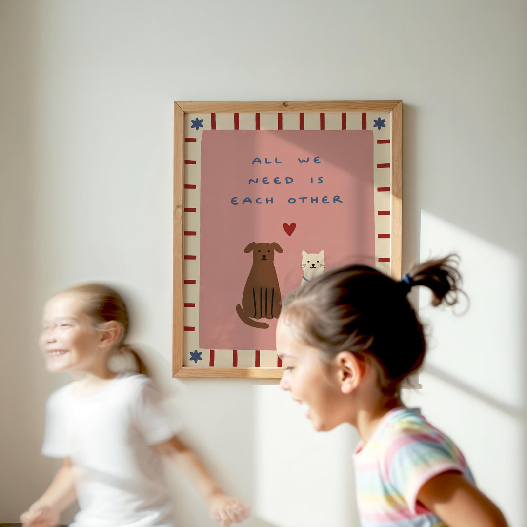 Two young girls are running in front of a framed poster with the text "All we need is each other" and illustrations of a dog and a cat.