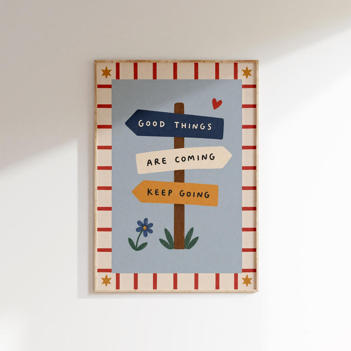 The image is a framed poster with a blue background and a red and white border. The poster features a sign with the text "Good Things Are Coming" and a heart symbol, along with a flower and a star.