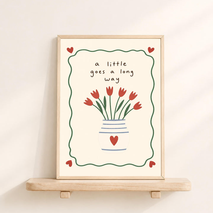 A framed illustration of a vase with red flowers and hearts, accompanied by the text "a little goes a long way" in a decorative border.