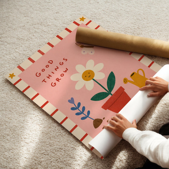 A child's hands are seen rolling up a poster with a colorful design of a flower, leaves, and a watering can.