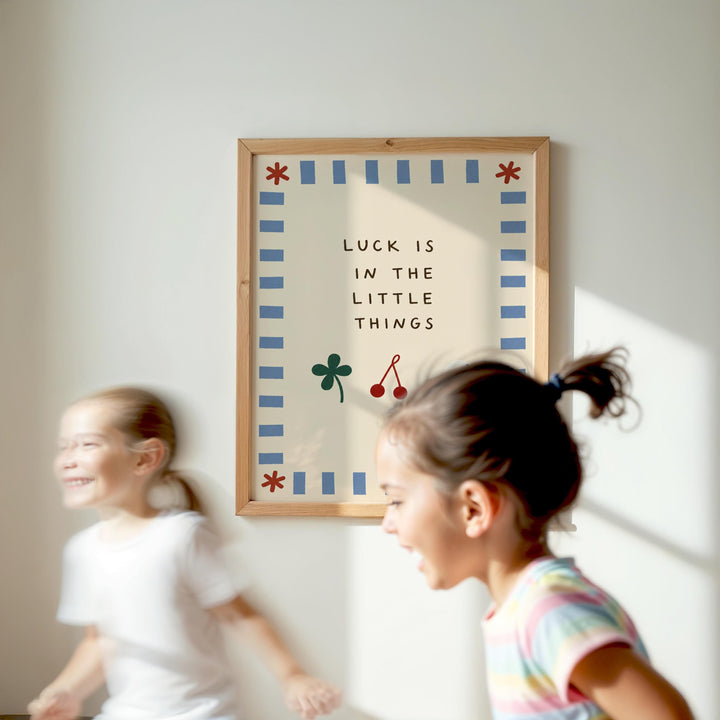 Two young girls are running in front of a framed sign with the text "LUCK IS IN THE LITTLE THINGS" and a cloverleaf design.