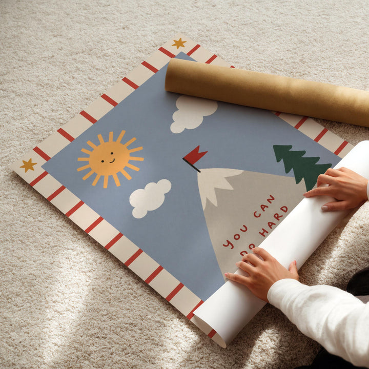 A child's hands are seen rolling up a poster with a mountain and sun design, which appears to be a colorful and playful artwork.