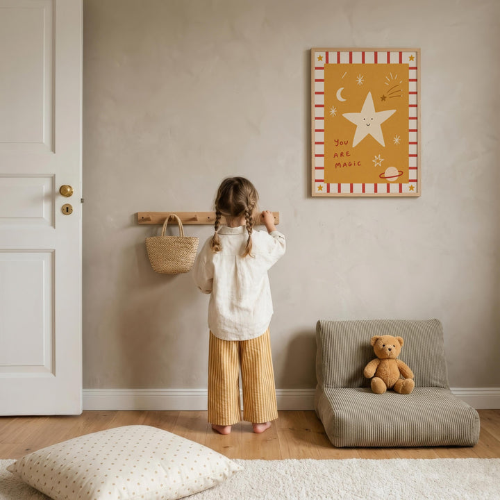 A young girl stands in a cozy room, looking up at a framed picture on the wall. The room has a wooden floor, a small chair, and a teddy bear on the floor.