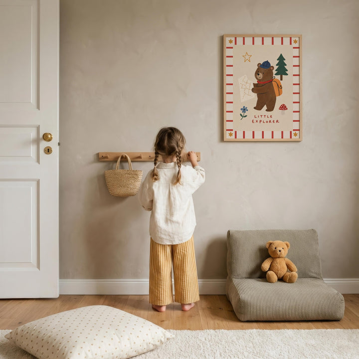A young girl stands in a cozy room, looking at a framed picture of a bear on the wall. The room has a beige color scheme, with a teddy bear sitting on a chair and a basket on a shelf.