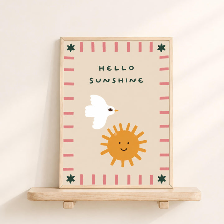 A framed illustration on a wooden shelf, depicting a white bird, a smiling sun, and a pink and green border.