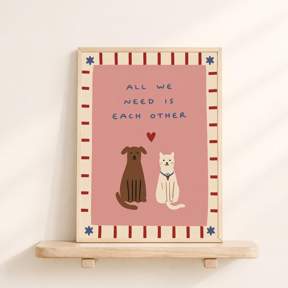 a framed illustration of a brown dog and a white cat sitting together, with the text "All we need is each other" written above them.