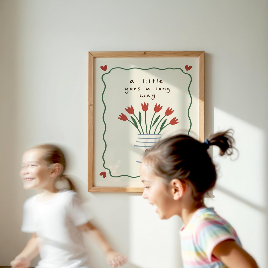 Two young girls are running in front of a framed picture with a vase of red flowers and the text "a little goes a long way" on the wall.