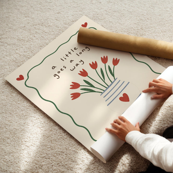 A person's hands are rolling up a poster with a floral design and the text "a little goes a long way" on it.