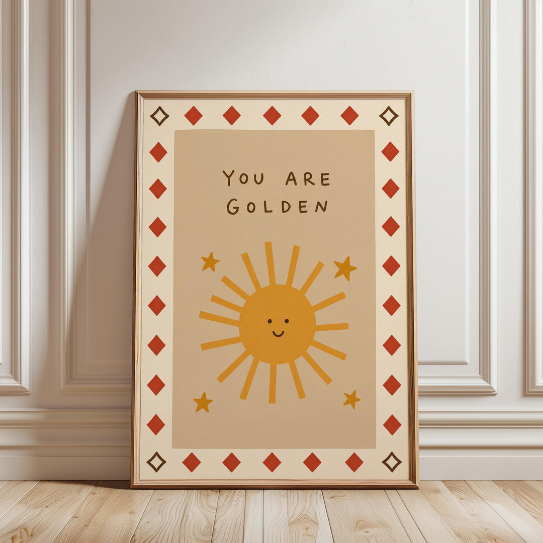 A framed poster with a cheerful sun design and the text "You are Golden" on it, placed on a wooden floor against a white wall.