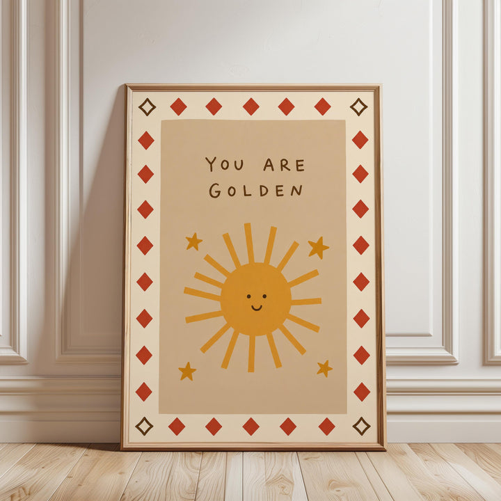 A framed poster with a cheerful sun design and the text "You are Golden" on it, placed on a wooden floor against a white wall.