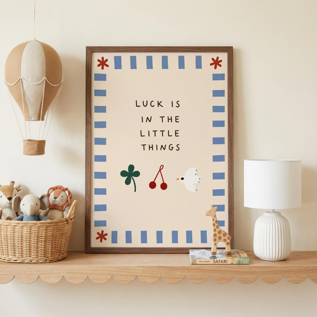 a framed poster with the text "LUCK IS IN THE LITTLE THINGS" and illustrations of a hot air balloon, a toy elephant, a toy giraffe, and a toy bird. The poster is displayed on a shelf next to a basket filled with stuffed animals and a white lamp.