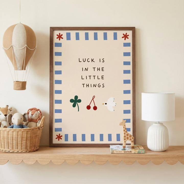 a framed poster with the text "LUCK IS IN THE LITTLE THINGS" and illustrations of a hot air balloon, a toy elephant, a toy giraffe, and a toy bird. The poster is displayed on a shelf next to a basket filled with stuffed animals and a white lamp.