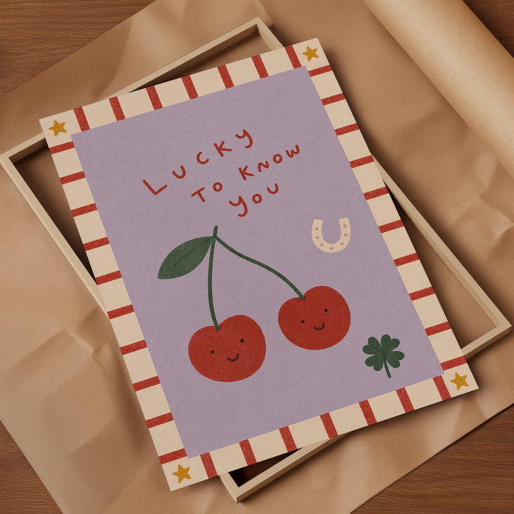 A greeting card with a purple background, featuring illustrations of cherries and a horseshoe. The text on the card reads "Lucky to know you".