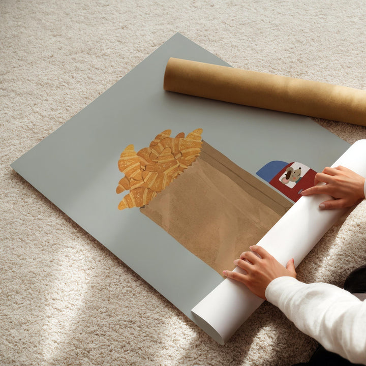 A person's hands are rolling up a roll of paper, which is placed on a gray surface. The paper has an illustration of a bag filled with golden-brown croissants.