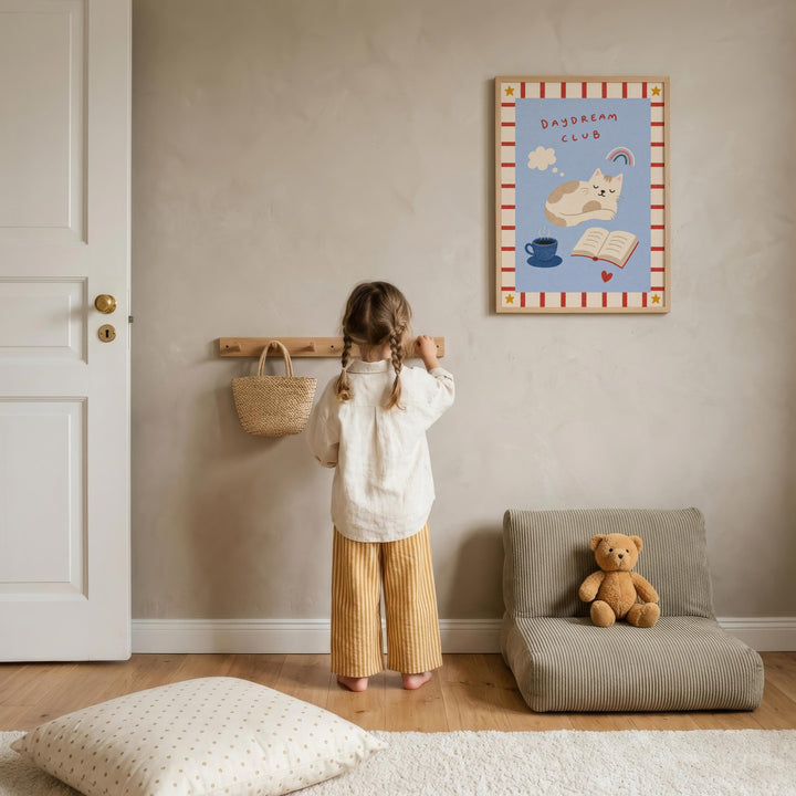 A young girl stands in a cozy room, looking at a framed poster on the wall. The poster features a cat and a cup, and the girl is wearing a white shirt and striped pants. A teddy bear is sitting on a small couch next to the girl, and a wooden shelf is visible on the wall.