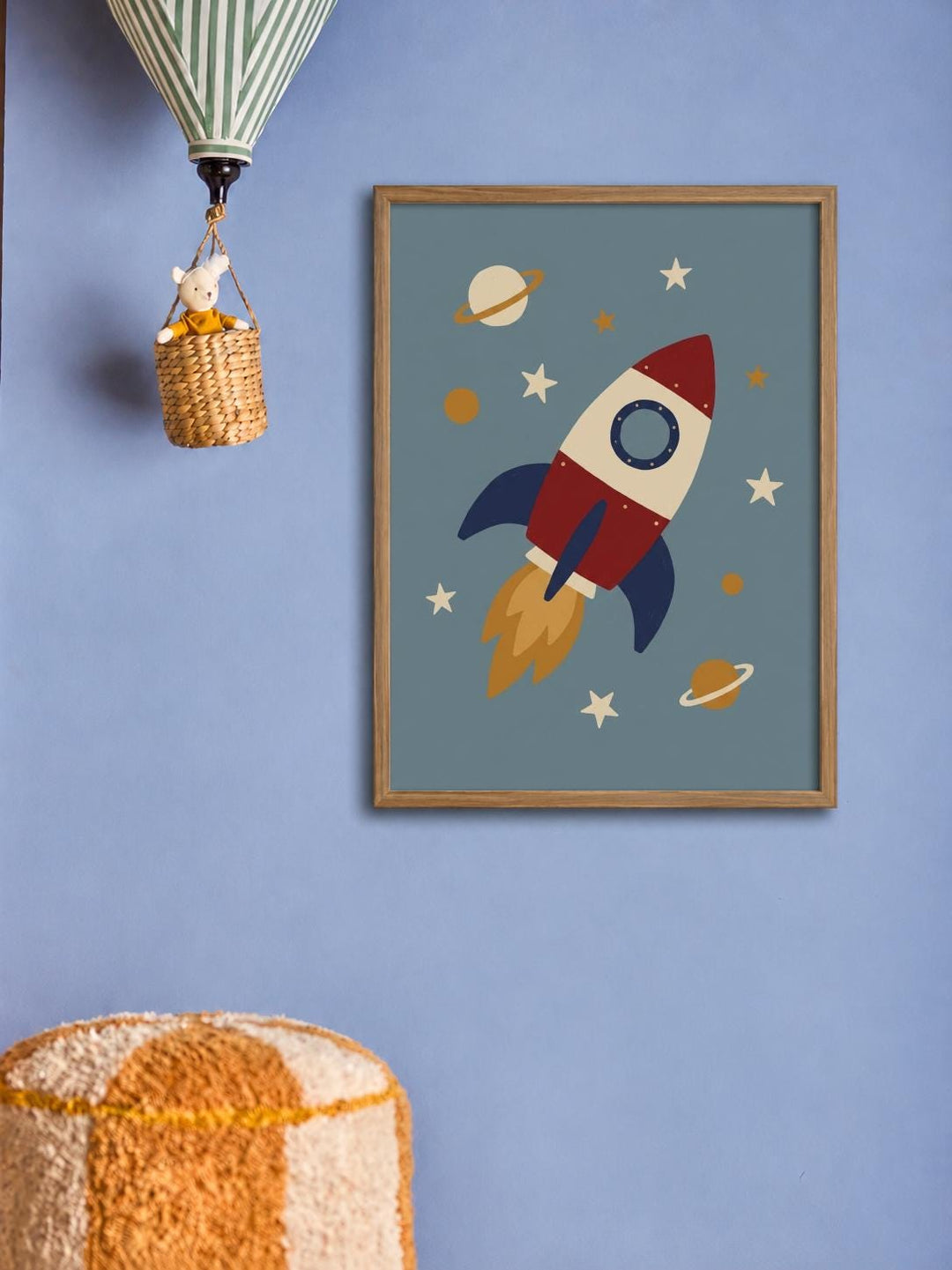 a framed picture of a rocket ship with planets and stars on a blue background, hanging on a wall. There is also a basket with a toy hanging from it and a hot air balloon in the background.