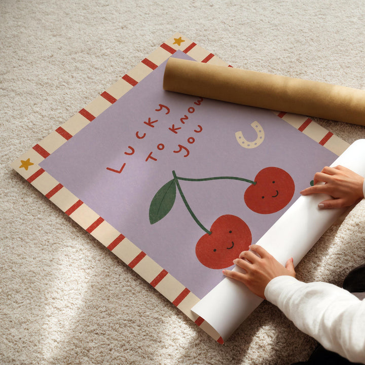 A person's hands are rolling up a purple poster with a cartoon image of cherries and the text "Lucky to know you" on it.