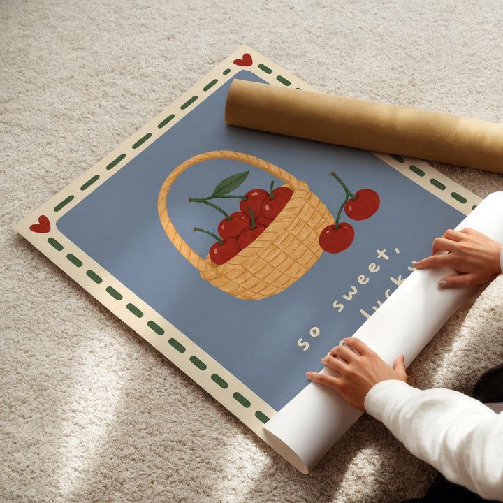 A person's hands are rolling up a poster featuring a basket of cherries and the text "So sweet, luck" on a carpeted floor.