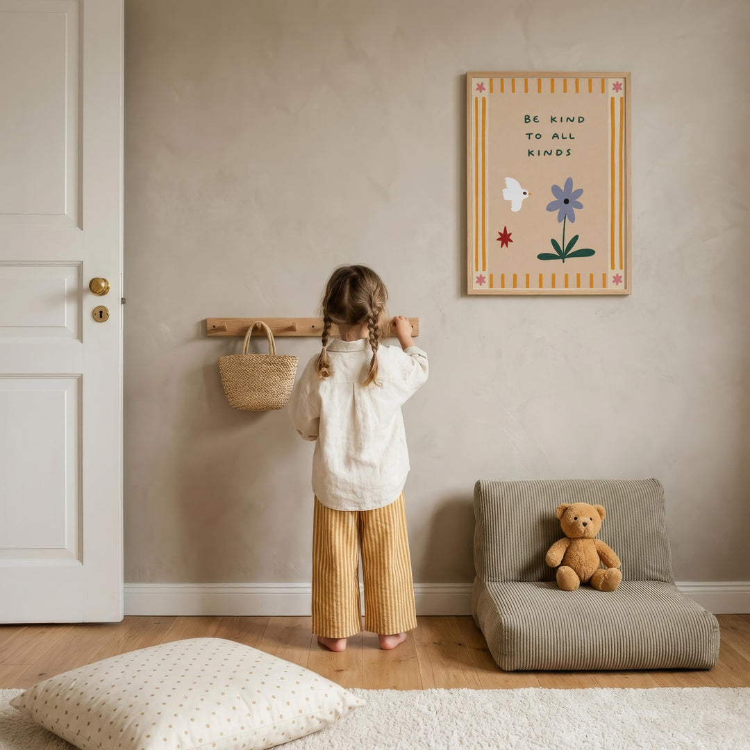 A young girl stands in a cozy room, looking at a wall-mounted sign that reads "Be kind to all kinds". The room has a beige color scheme, with a teddy bear sitting on a small couch and a wooden shelf with a woven basket nearby.