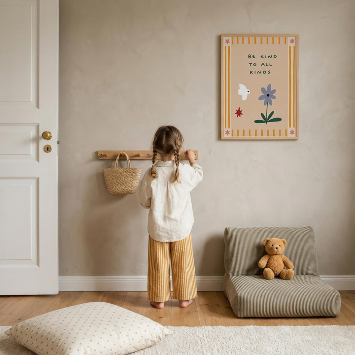 A young girl stands in a cozy room, looking at a wall-mounted sign that reads "Be kind to all kinds". The room has a beige color scheme, with a teddy bear sitting on a small couch and a wooden shelf with a woven basket nearby.