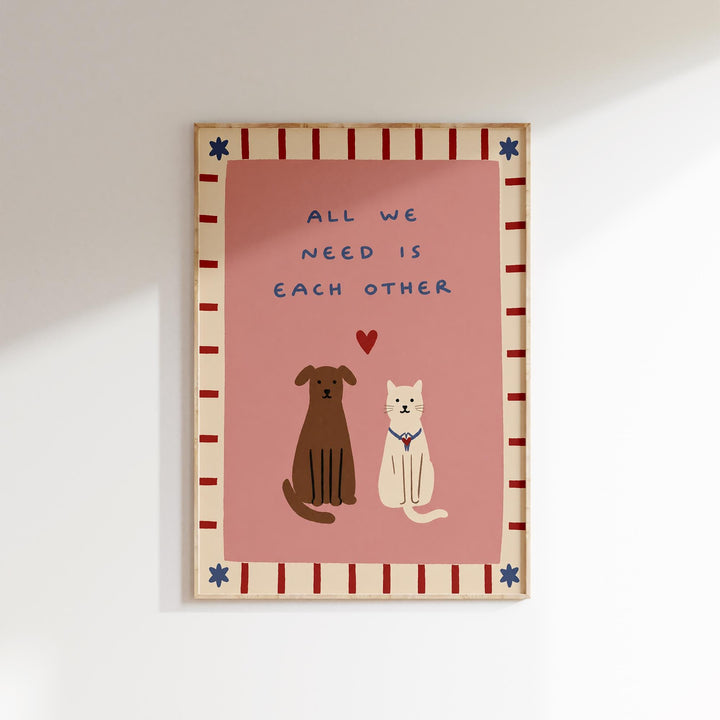 a framed illustration of two dogs and a cat sitting together, with a heart symbol between them, and the text "All we need is each other" written above them.