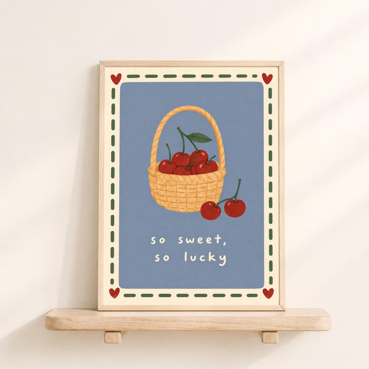 A framed illustration of a basket filled with cherries, accompanied by the text "so sweet, so lucky" in a decorative border.