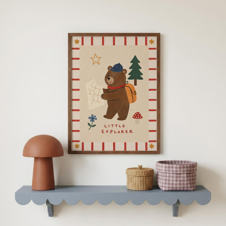 a framed poster of a bear holding a map and a mushroom, with the text "Little Explorer" below the bear. The poster is displayed on a wall above a shelf with a mushroom-shaped lamp and a woven basket.