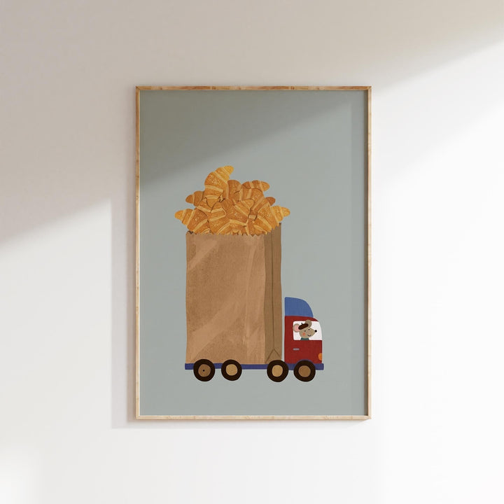 A framed illustration depicting a large brown paper bag filled with golden-brown croissants, with a red truck carrying the bag.